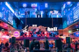 A vibrant and bustling urban scene featuring an illuminated facade with neon signs and advertisements. People are gathered in a lively atmosphere, with some individuals standing on an upper level observing the scene below. The area is filled with colorful lights and dynamic displays.