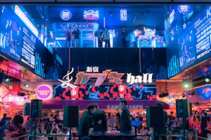 A vibrant and bustling urban scene featuring an illuminated facade with neon signs and advertisements. People are gathered in a lively atmosphere, with some individuals standing on an upper level observing the scene below. The area is filled with colorful lights and dynamic displays.