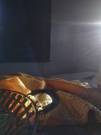 Sunlight streams into a dimly lit room, illuminating a table adorned with a yellow cloth and a mesh-like fabric. A window with a dark, rolled-down shade is in the background, and a decorative metal object with intricate patterns is placed in the foreground. The sunlight creates a warm and serene atmosphere with light rays visibly spreading across the scene.