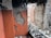 An interior space with exposed brick walls and visible structural damage. The plaster is crumbling, revealing bricks beneath, and there is a pile of rubble on the floor. The walls have patches of orange paint, and some bricks are missing, indicating significant deterioration.