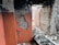 An interior space with exposed brick walls and visible structural damage. The plaster is crumbling, revealing bricks beneath, and there is a pile of rubble on the floor. The walls have patches of orange paint, and some bricks are missing, indicating significant deterioration.