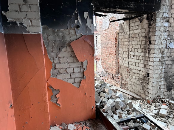 An interior space with exposed brick walls and visible structural damage. The plaster is crumbling, revealing bricks beneath, and there is a pile of rubble on the floor. The walls have patches of orange paint, and some bricks are missing, indicating significant deterioration.