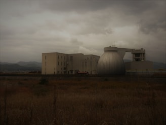 A large industrial building complex under overcast skies, featuring a distinctive spherical structure adjacent to several rectangular buildings. In the foreground, dry grass stretches across a field with mountains faintly visible in the background.