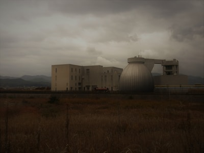 A large industrial building complex under overcast skies, featuring a distinctive spherical structure adjacent to several rectangular buildings. In the foreground, dry grass stretches across a field with mountains faintly visible in the background.
