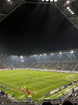 A nighttime soccer match in a large, well-lit stadium. Players in brightly colored jerseys are on the field, surrounded by a large audience in the stands. The scoreboard displays 0-0, indicating the start of the match. The seating area features patterns made by colored seats among the spectators.