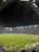 A nighttime soccer match in a large, well-lit stadium. Players in brightly colored jerseys are on the field, surrounded by a large audience in the stands. The scoreboard displays 0-0, indicating the start of the match. The seating area features patterns made by colored seats among the spectators.