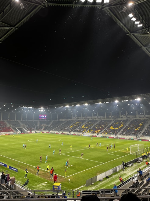 A nighttime soccer match in a large, well-lit stadium. Players in brightly colored jerseys are on the field, surrounded by a large audience in the stands. The scoreboard displays 0-0, indicating the start of the match. The seating area features patterns made by colored seats among the spectators.