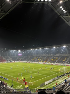 A nighttime soccer match in a large, well-lit stadium. Players in brightly colored jerseys are on the field, surrounded by a large audience in the stands. The scoreboard displays 0-0, indicating the start of the match. The seating area features patterns made by colored seats among the spectators.