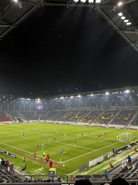A nighttime soccer match in a large, well-lit stadium. Players in brightly colored jerseys are on the field, surrounded by a large audience in the stands. The scoreboard displays 0-0, indicating the start of the match. The seating area features patterns made by colored seats among the spectators.