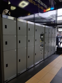 A row of gray lockers with numbered doors is arranged along a corridor. The lockers have black handles and are situated indoors, with part of the ceiling and floor visible. There is a security camera mounted on the upper left and an advertisement banner above.