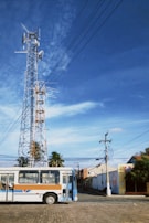 A tall communication tower with multiple antennas and dishes stands against a clear blue sky. Below, a bus with a brown, white, and blue design is parked on a cobblestone road. Surrounding the scene are palm trees and low-rise buildings with utility poles connected by wires.