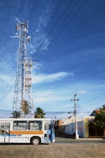 A tall communication tower with multiple antennas and dishes stands against a clear blue sky. Below, a bus with a brown, white, and blue design is parked on a cobblestone road. Surrounding the scene are palm trees and low-rise buildings with utility poles connected by wires.