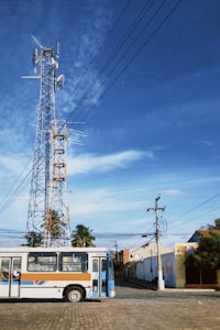 A tall communication tower with multiple antennas and dishes stands against a clear blue sky. Below, a bus with a brown, white, and blue design is parked on a cobblestone road. Surrounding the scene are palm trees and low-rise buildings with utility poles connected by wires.