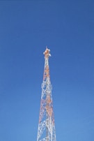 A tall communication tower with an orange and white lattice structure reaches up into a clear blue sky. The tower is equipped with several antennas.