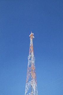A tall communication tower with an orange and white lattice structure reaches up into a clear blue sky. The tower is equipped with several antennas.