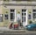 Two people walking past a storefront with green walls and white doors, featuring art and jewelry posters. There's a red scooter parked on the sidewalk and a blue car on the street. The scene includes some graffiti on the wall and a small potted plant near the entrance.