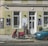Two people walking past a storefront with green walls and white doors, featuring art and jewelry posters. There's a red scooter parked on the sidewalk and a blue car on the street. The scene includes some graffiti on the wall and a small potted plant near the entrance.