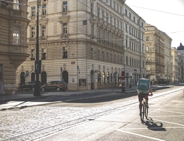 A cyclist rides down a quiet, sunlit street lined with ornate, historic buildings. The cyclist carries a turquoise delivery backpack, and the cobblestone street reflects the early morning or late afternoon sun. There are a few parked cars and empty sidewalks, suggesting a calm, urban setting.