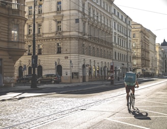 A cyclist rides down a quiet, sunlit street lined with ornate, historic buildings. The cyclist carries a turquoise delivery backpack, and the cobblestone street reflects the early morning or late afternoon sun. There are a few parked cars and empty sidewalks, suggesting a calm, urban setting.