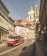 A vintage red and cream tram travels along a cobblestone street lined with historic European buildings. The street is well-lit by natural sunlight, and the tram cables crisscross the sky. In the background, an ornate church with a prominent dome and spire adds to the classic cityscape scenery.