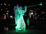 A luminous sculpture of an angel made from wireframe and lights, playing a trumpet. The sculpture emits a bright green glow, standing in contrast to the dark night background. Other decorative lights are visible in the distance, adding to the festive atmosphere.