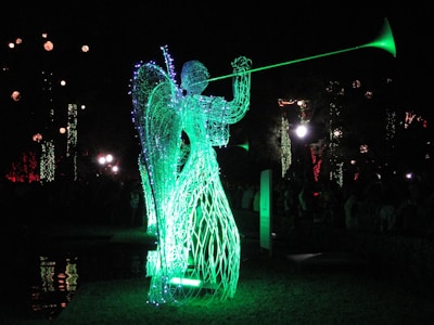 A luminous sculpture of an angel made from wireframe and lights, playing a trumpet. The sculpture emits a bright green glow, standing in contrast to the dark night background. Other decorative lights are visible in the distance, adding to the festive atmosphere.
