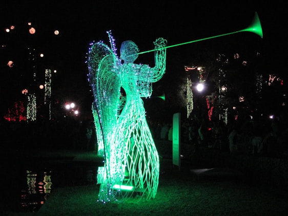 A luminous sculpture of an angel made from wireframe and lights, playing a trumpet. The sculpture emits a bright green glow, standing in contrast to the dark night background. Other decorative lights are visible in the distance, adding to the festive atmosphere.