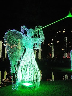 A glowing wire-frame sculpture of an angel playing a trumpet, illuminated in bright green and purple lights. The angel stands on grass near a body of water, with reflections of the lights visible in the water. In the background, numerous circular lights and light-adorned trees enhance the magical and festive atmosphere.