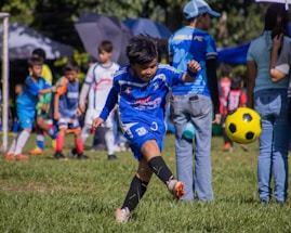 A young boy is playing soccer on a grassy field, dressed in a blue soccer uniform. He is in mid-kick, focused on the yellow and black soccer ball in front of him. Several other children wearing soccer uniforms are in the background, some standing and some playing. Adults are also present, possibly supervising the activity. There is a mix of bright and shadowed areas in the setting.