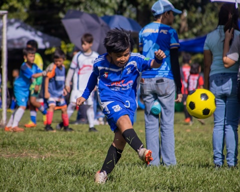 A young boy is playing soccer on a grassy field, dressed in a blue soccer uniform. He is in mid-kick, focused on the yellow and black soccer ball in front of him. Several other children wearing soccer uniforms are in the background, some standing and some playing. Adults are also present, possibly supervising the activity. There is a mix of bright and shadowed areas in the setting.