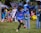 A young boy is playing soccer on a grassy field, dressed in a blue soccer uniform. He is in mid-kick, focused on the yellow and black soccer ball in front of him. Several other children wearing soccer uniforms are in the background, some standing and some playing. Adults are also present, possibly supervising the activity. There is a mix of bright and shadowed areas in the setting.