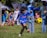A young boy is playing soccer on a grassy field, dressed in a blue soccer uniform. He is in mid-kick, focused on the yellow and black soccer ball in front of him. Several other children wearing soccer uniforms are in the background, some standing and some playing. Adults are also present, possibly supervising the activity. There is a mix of bright and shadowed areas in the setting.