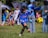 A young boy is playing soccer on a grassy field, dressed in a blue soccer uniform. He is in mid-kick, focused on the yellow and black soccer ball in front of him. Several other children wearing soccer uniforms are in the background, some standing and some playing. Adults are also present, possibly supervising the activity. There is a mix of bright and shadowed areas in the setting.
