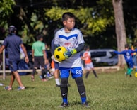 A young boy in a white and blue soccer uniform, wearing goalkeeper gloves, is holding a yellow and black soccer ball on a grassy field. Other children and a few adults can be seen in the background, some wearing sports attire, indicating a soccer practice or game in progress. Trees and parked cars are visible in the background, providing a park-like setting.