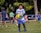 A young boy in a white and blue soccer uniform, wearing goalkeeper gloves, is holding a yellow and black soccer ball on a grassy field. Other children and a few adults can be seen in the background, some wearing sports attire, indicating a soccer practice or game in progress. Trees and parked cars are visible in the background, providing a park-like setting.