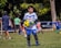 A young boy in a white and blue soccer uniform, wearing goalkeeper gloves, is holding a yellow and black soccer ball on a grassy field. Other children and a few adults can be seen in the background, some wearing sports attire, indicating a soccer practice or game in progress. Trees and parked cars are visible in the background, providing a park-like setting.
