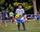 A young boy in a white and blue soccer uniform, wearing goalkeeper gloves, is holding a yellow and black soccer ball on a grassy field. Other children and a few adults can be seen in the background, some wearing sports attire, indicating a soccer practice or game in progress. Trees and parked cars are visible in the background, providing a park-like setting.