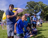 A group of children and adults are gathered at a soccer field, watching an event. The children are wearing blue sports uniforms and seem excited and cheerful. Some people hold umbrellas for shade, while others are standing near a goalpost in the background. The atmosphere appears lively and joyful, with a mix of natural and bright colors present.