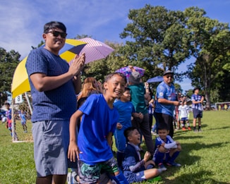 A group of children and adults are gathered at a soccer field, watching an event. The children are wearing blue sports uniforms and seem excited and cheerful. Some people hold umbrellas for shade, while others are standing near a goalpost in the background. The atmosphere appears lively and joyful, with a mix of natural and bright colors present.