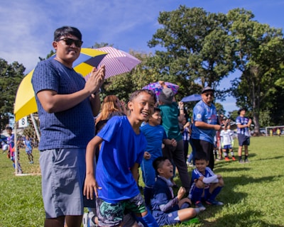 A group of children and adults are gathered at a soccer field, watching an event. The children are wearing blue sports uniforms and seem excited and cheerful. Some people hold umbrellas for shade, while others are standing near a goalpost in the background. The atmosphere appears lively and joyful, with a mix of natural and bright colors present.