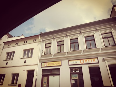 Two adjacent buildings with white facades are visible, featuring large windows and signs indicating an Asian bistro and an optical store. The architectural style includes simple, classical elements with overhanging eaves and decorative window frames. The sky is partly cloudy, creating a soft, diffused lighting that casts mild shadows.