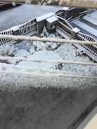 A snowy scene with accumulated snow on the ground, fence, and a series of electrical wires. The area is a fenced yard with minimal landscaping visible under the snow. Snow has evenly covered the pavement and wooden fence.