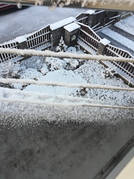 A snowy scene with accumulated snow on the ground, fence, and a series of electrical wires. The area is a fenced yard with minimal landscaping visible under the snow. Snow has evenly covered the pavement and wooden fence.