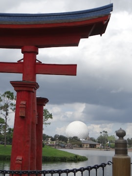 A large red Japanese torii gate stands prominently in the foreground, with a partly cloudy sky providing contrast. In the background, the iconic geodesic sphere, known as Spaceship Earth, is situated in a landscaped park area near a body of water.