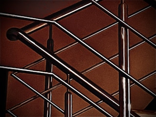 A close-up of a modern metal railing structure, featuring intersecting horizontal and vertical bars. The metallic elements contrast against a warm, brownish background, highlighting the sleek and polished nature of the metal.