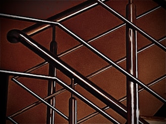 A close-up of a modern metal railing structure, featuring intersecting horizontal and vertical bars. The metallic elements contrast against a warm, brownish background, highlighting the sleek and polished nature of the metal.