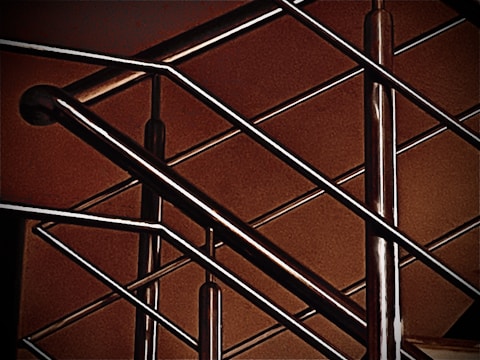 A close-up of a modern metal railing structure, featuring intersecting horizontal and vertical bars. The metallic elements contrast against a warm, brownish background, highlighting the sleek and polished nature of the metal.