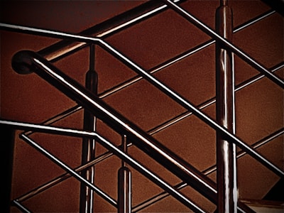 A close-up of a modern metal railing structure, featuring intersecting horizontal and vertical bars. The metallic elements contrast against a warm, brownish background, highlighting the sleek and polished nature of the metal.