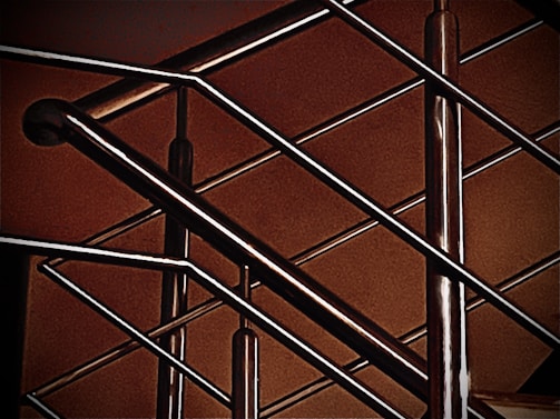 A close-up of a modern metal railing structure, featuring intersecting horizontal and vertical bars. The metallic elements contrast against a warm, brownish background, highlighting the sleek and polished nature of the metal.