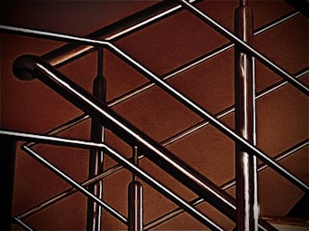 A close-up of a modern metal railing structure, featuring intersecting horizontal and vertical bars. The metallic elements contrast against a warm, brownish background, highlighting the sleek and polished nature of the metal.
